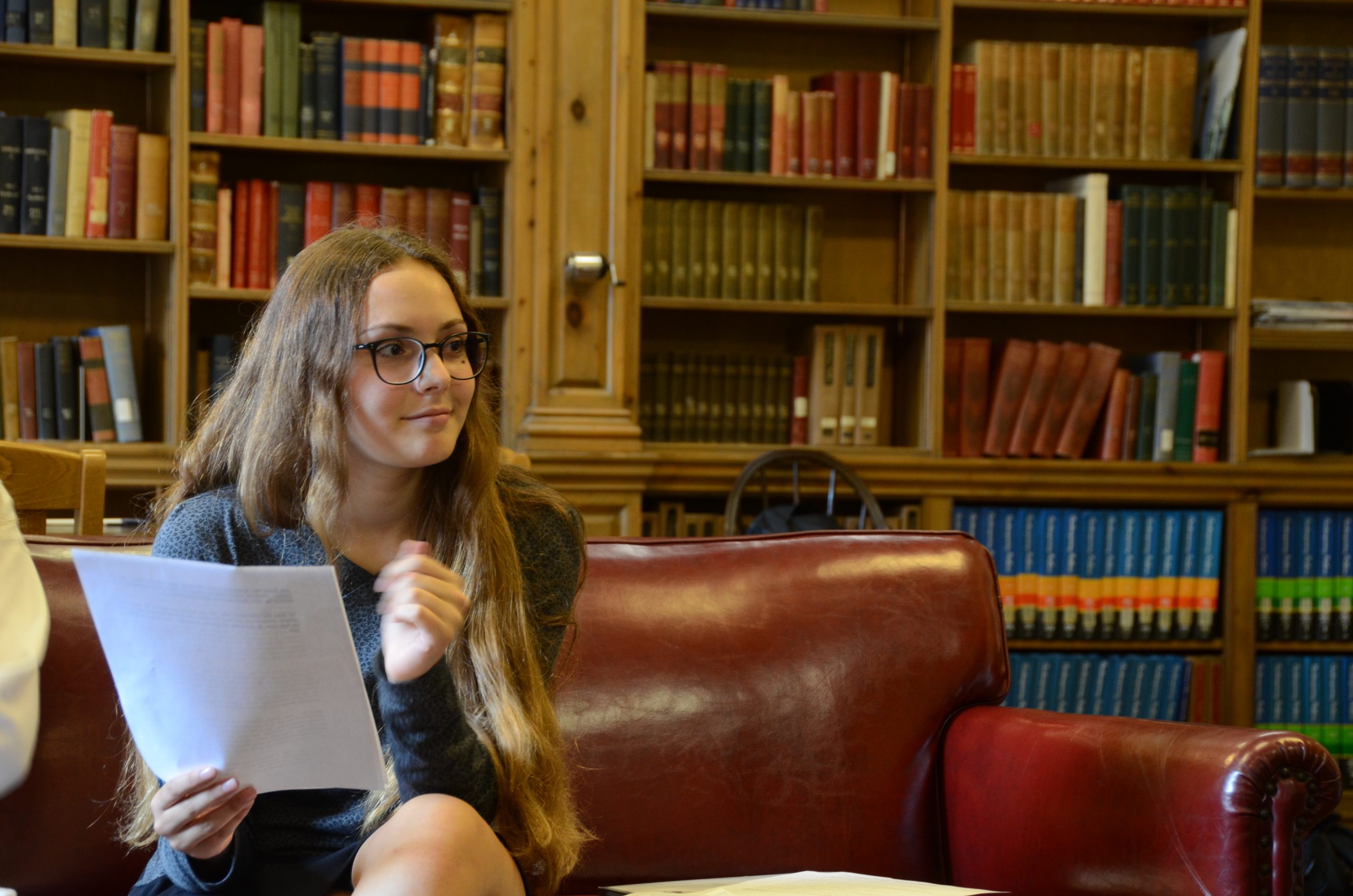 A person with long hair and glasses holds a document while sitting on a red leather sofa in a library. Shelves of colorful books are visible in the background.