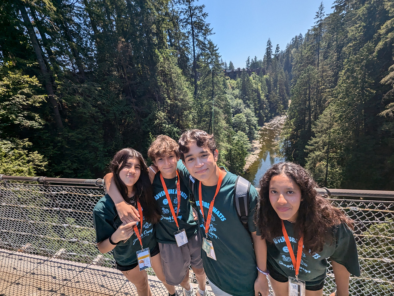 Four teenagers wearing camp shirts and lanyards stand together on a bridge surrounded by tall trees and a river below, on a sunny day.