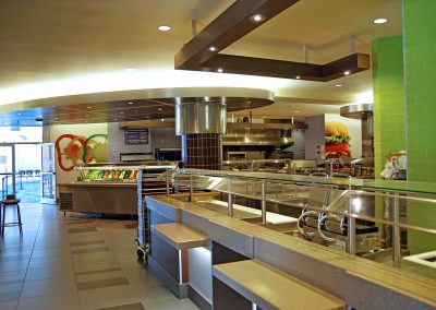 A modern cafeteria interior with food service counters, salad bar, stainless steel equipment, and seating areas. Large food images decorate the wall in the background.