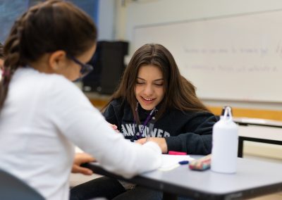 Two students sit at a classroom table, one writing on paper while the other watches. A whiteboard and water bottle are visible in the background.