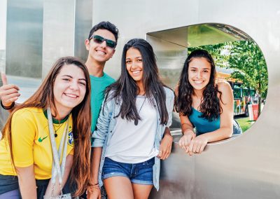 Four young people pose and smile outdoors next to a large metal structure on a sunny day; one person wears a yellow Brazil shirt.
