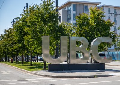 Large metallic letters spelling "UBC" stand by a sidewalk next to trees and a modern building on a sunny day.