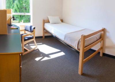 A small dorm room with a single bed, a desk and chair, and a window letting in natural light.