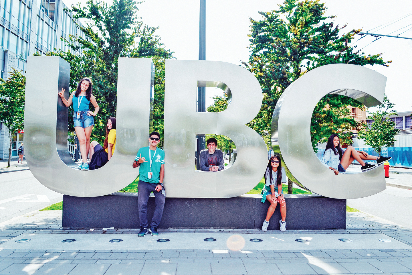 UBC Six young adults pose and sit around large metallic letters spelling "UBC" outdoors on a sunny day, with trees and buildings in the background.