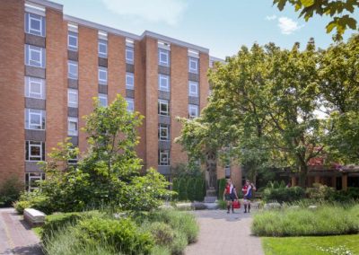 A five-story brick dormitory building with several windows, surrounded by green trees and bushes; two people walk along a path in the foreground.