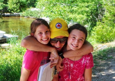 An adult wearing a yellow hat hugs two smiling children outdoors near a lake, with trees and a dock in the background.
