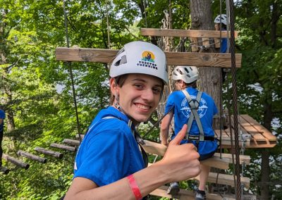 A person wearing a helmet and harness gives a thumbs up while sitting on a wooden platform in a forest ropes course. Other participants are visible in the background.