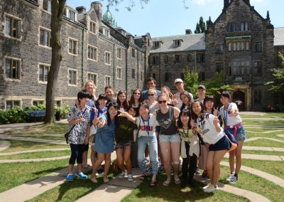 A group of young people pose and smile for a photo in a courtyard surrounded by historic stone buildings on a sunny day.