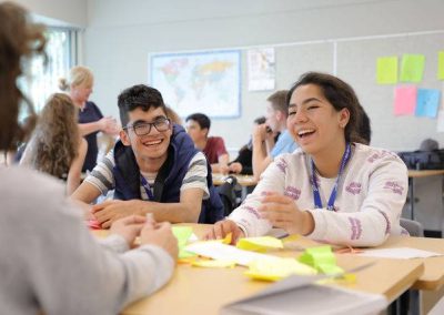 Students sit around a classroom table, smiling and talking, with colorful papers and classroom decorations visible in the background.