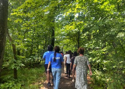 A group of people, including two in blue "STAFF" shirts, walk on a wooded trail surrounded by green trees on a sunny day.
