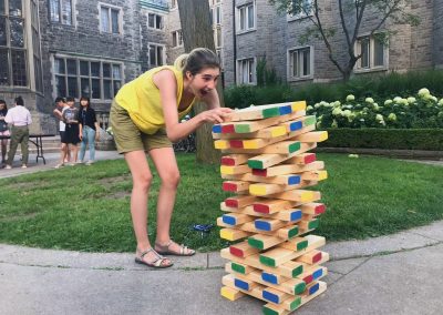 A woman in a yellow top plays a large outdoor Jenga game on a lawn with old stone buildings in the background.