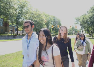 A group of young adults walk together outside on a sunny day, smiling, with trees and traffic lights visible in the background.