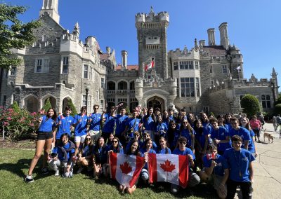 A large group of young people in blue shirts pose with Canada flags in front of a historic stone castle on a sunny day.
