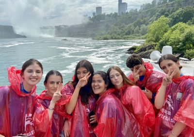 A group of six young people in red ponchos pose and smile near a body of water with Niagara Falls and city buildings visible in the background.