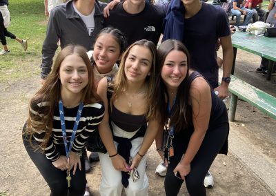 Six young people pose and smile for a photo in a park, with trees and a green picnic table visible in the background.