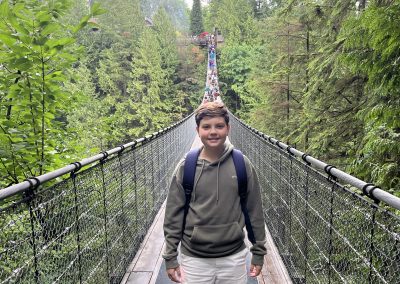 A boy wearing a green hoodie, white shorts, and a backpack stands on a suspension bridge surrounded by tall green trees.