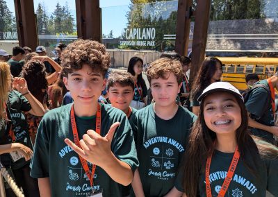 A group of children in matching green camp shirts stand together outside Capilano Suspension Bridge Park, with a school bus and other kids in the background.
