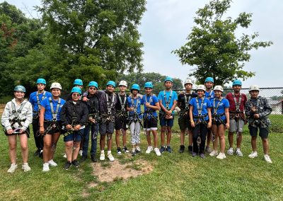 A group of people wearing helmets and harnesses stand together outdoors on grass, preparing for a zip-lining or ropes course activity. Trees and a fence are in the background.