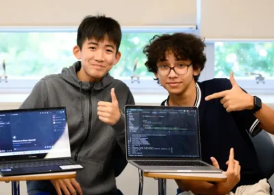 Two students sitting at desks with open laptops, one displaying a chat screen and the other showing code; both students are smiling and gesturing toward the laptops.