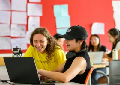 Two students sit at a desk working together on a laptop, smiling, in a classroom with sketches and drawings on a red wall in the background.