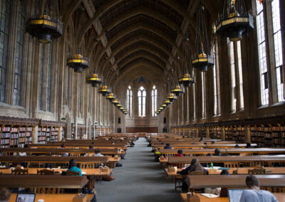 Large, gothic-style library reading room with arched ceiling, tall windows, hanging lights, rows of wooden tables, and people studying.