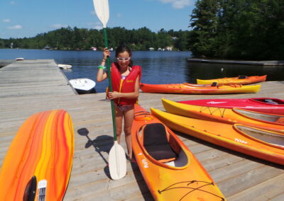 A girl in a red life jacket stands on a dock holding a paddle, next to several orange kayaks, with a lake and trees in the background.