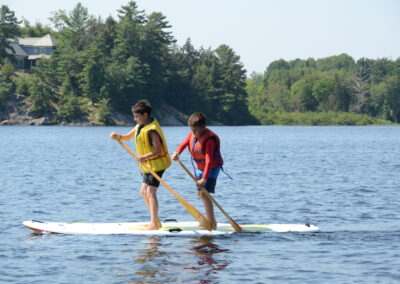 Two children stand on a paddleboard wearing life jackets, paddling together on a calm lake with trees and houses in the background.