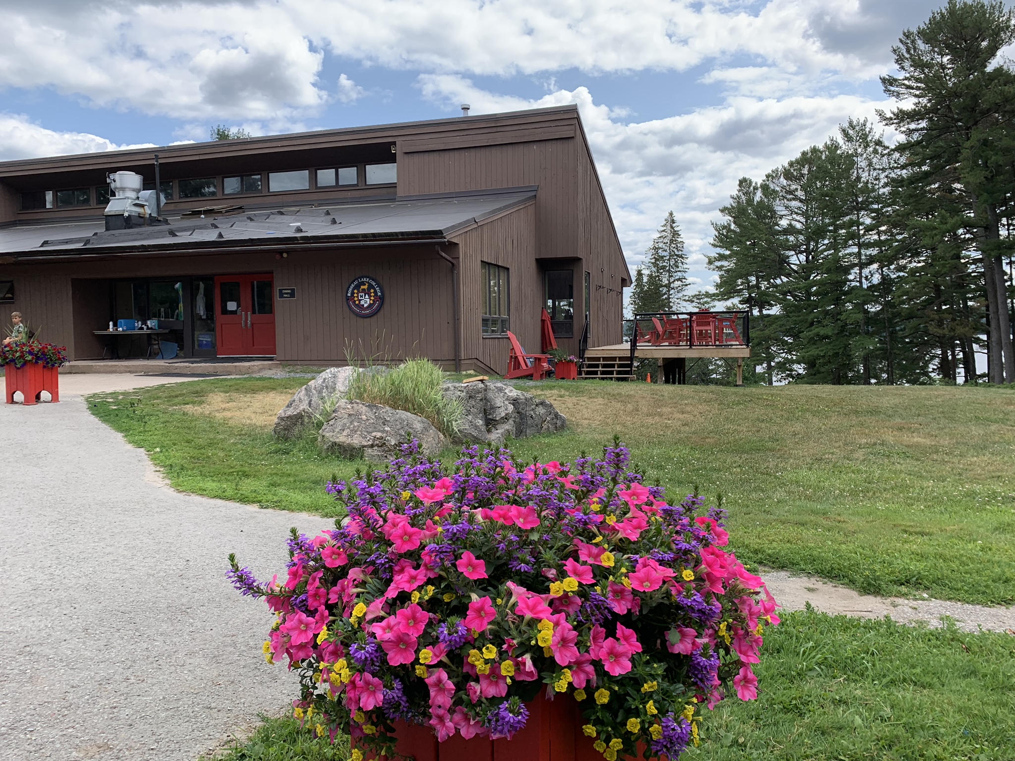 Brown building with red doors and windows, surrounded by green grass, trees, and a large planter filled with pink and yellow flowers in the foreground under a partly cloudy sky.