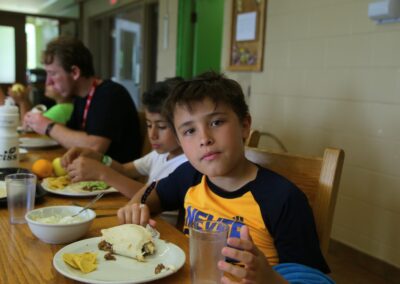 A boy sits at a table eating a meal with other children, holding a glass of water and looking at the camera. Food including a burrito and chips is on his plate.