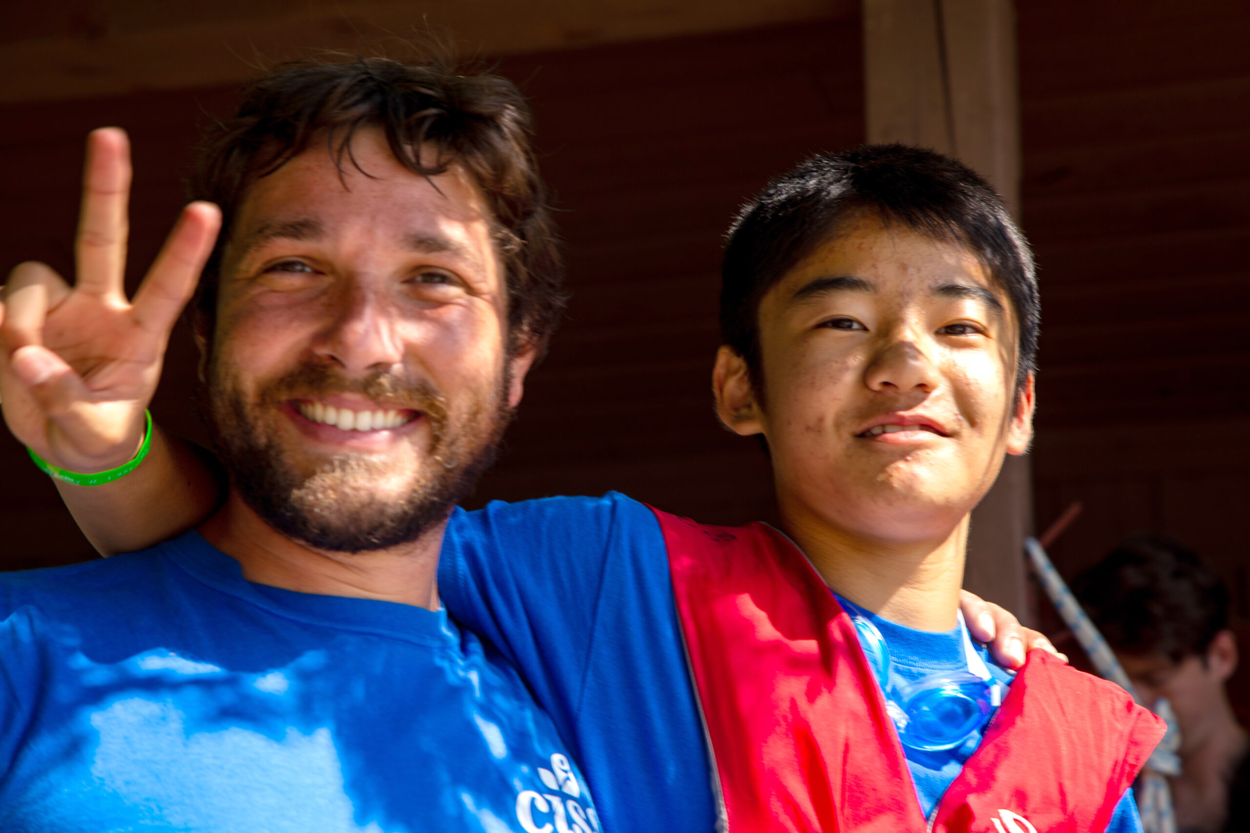 A man with a beard making a peace sign with his fingers has his arm around the shoulder of a teenage boy.